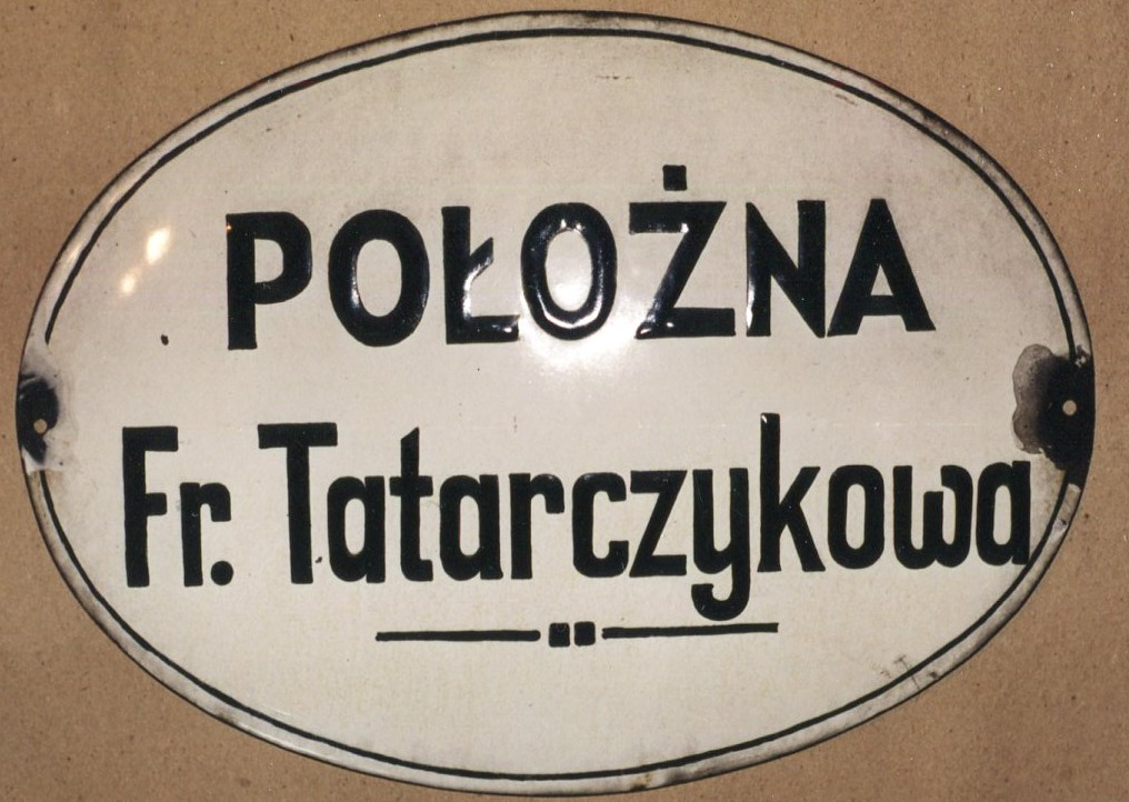 Tablica emaliowana, owalna. Na białym tle czarny napis: Położna Fr. Tatarczykowa